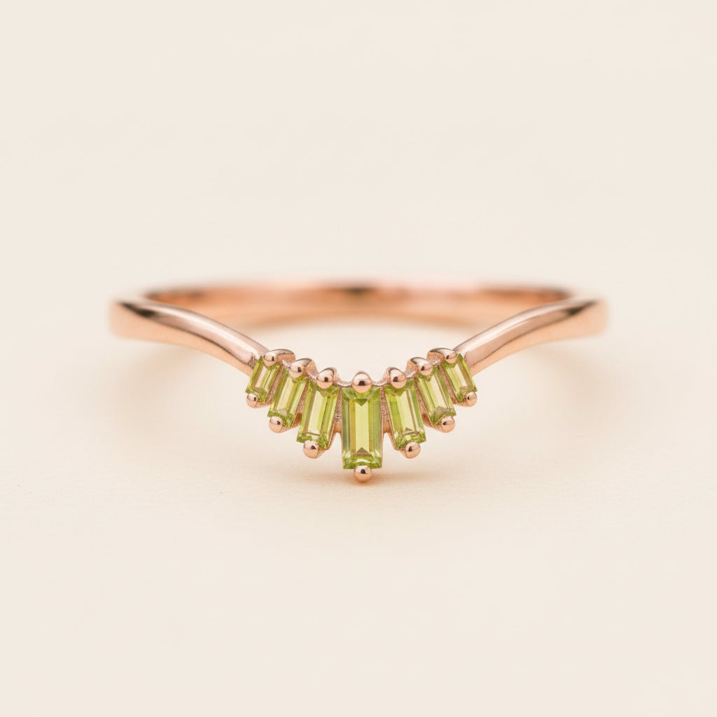 A rose gold ring with a curved band and a row of baguette-cut green peridot gemstones, resting on a textured stone surface with a blurred natural background.
