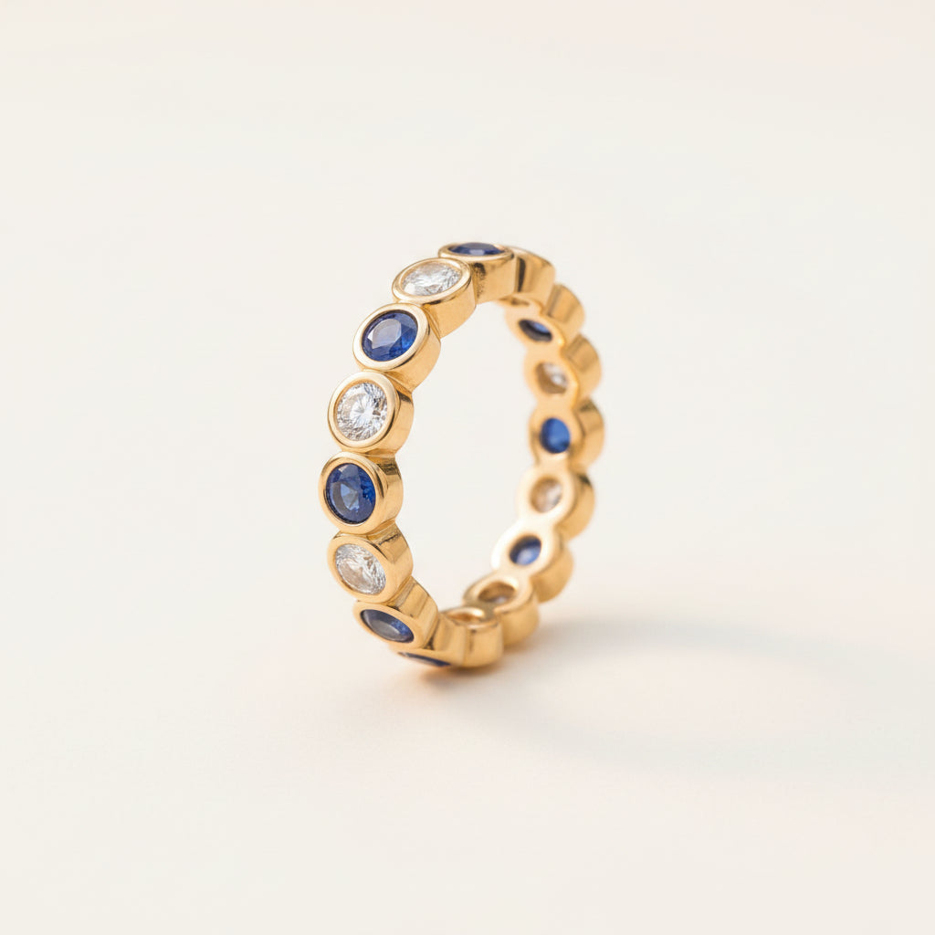 A gold eternity ring featuring alternating round blue sapphires and clear round diamonds, resting on a textured stone surface with a soft, blurred natural background.