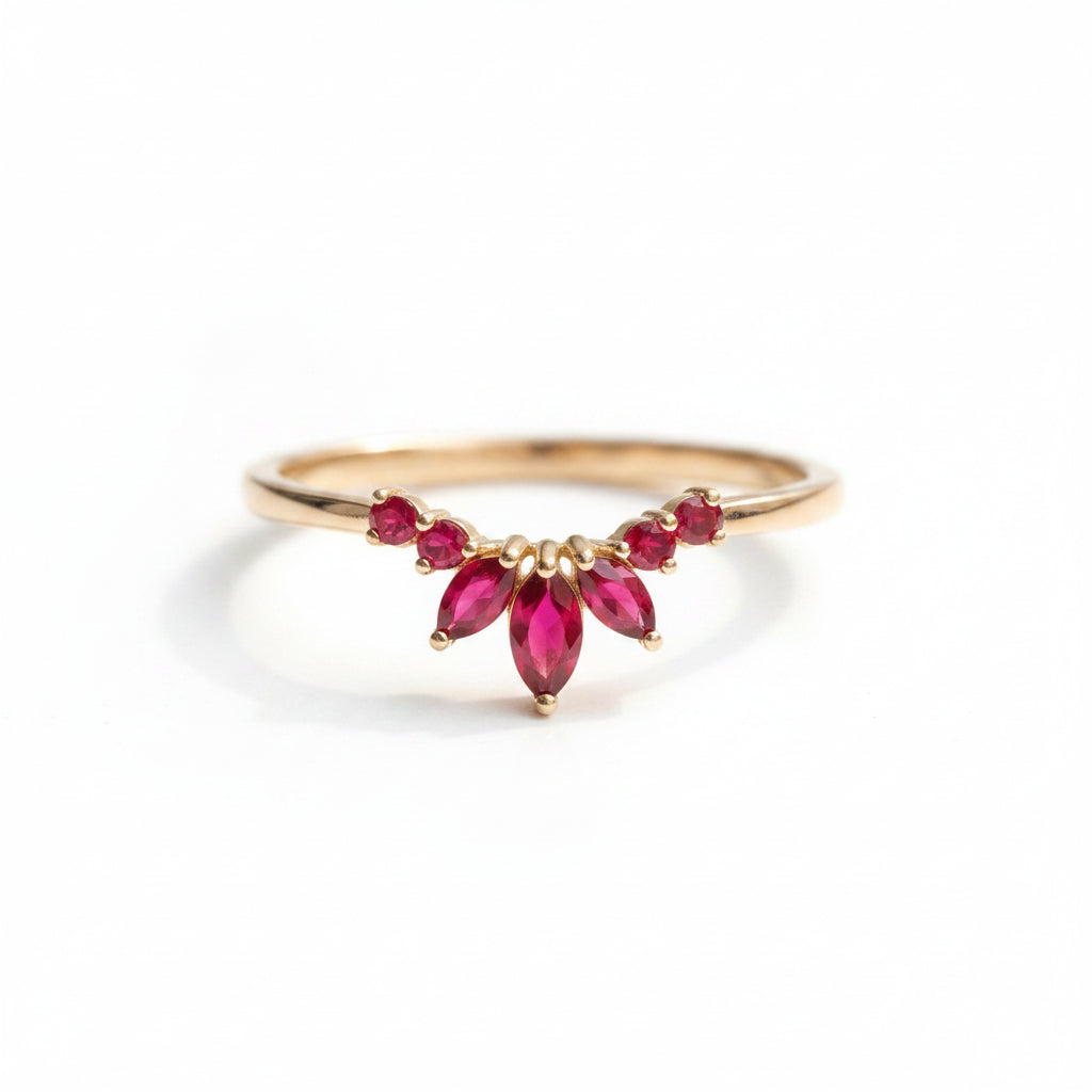 A gold ring featuring a curved row of red marquise and round gemstones, resting on a textured cream lace fabric with soft green leaves blurred in the background.