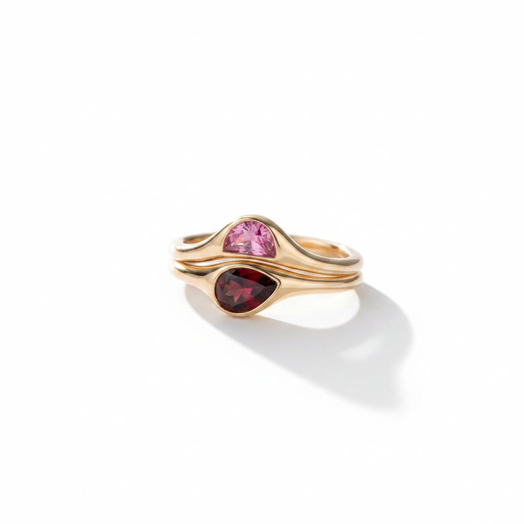 A gold ring with two teardrop-shaped gemstones, one pink and one deep red, displayed on a dark slate surface.