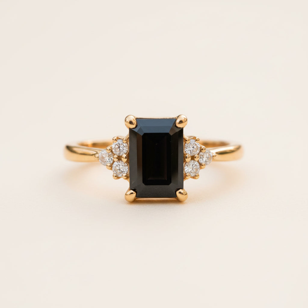 A yellow gold ring featuring a large rectangular black gemstone center with small clear accent stones on either side, resting on a textured stone surface with a blurred natural background.