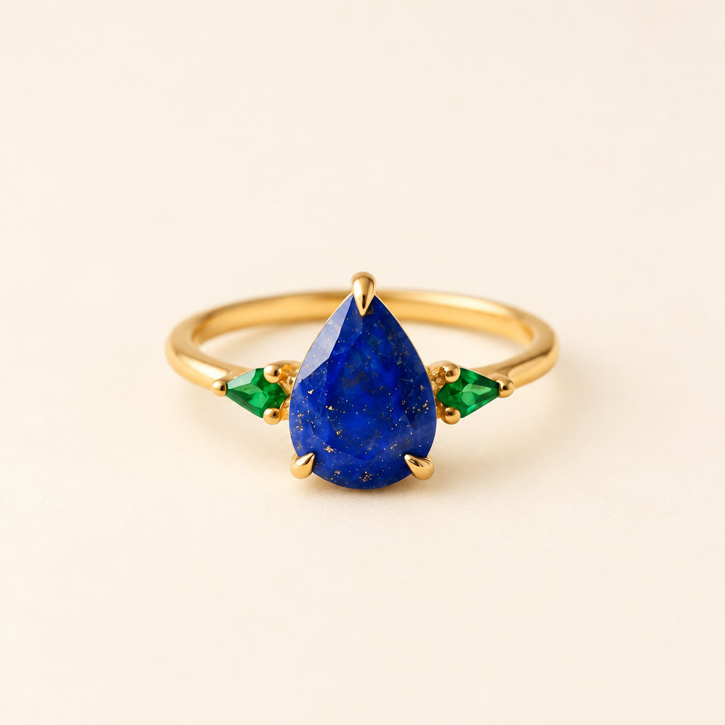 Gold ring with a blue teardrop gemstone and green accents on a textured surface with flowers.