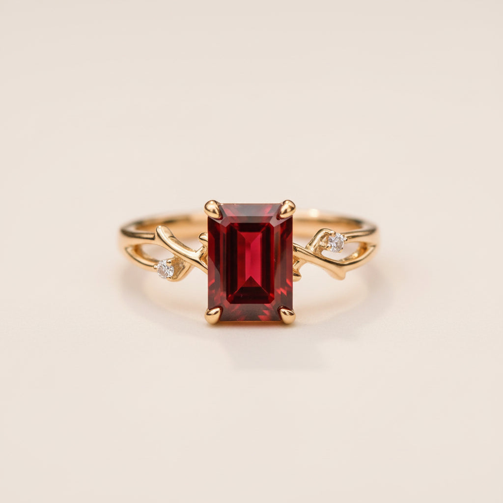 A gold ring with a large rectangular red gemstone and small side stones, resting on a textured stone surface with a blurred natural background.