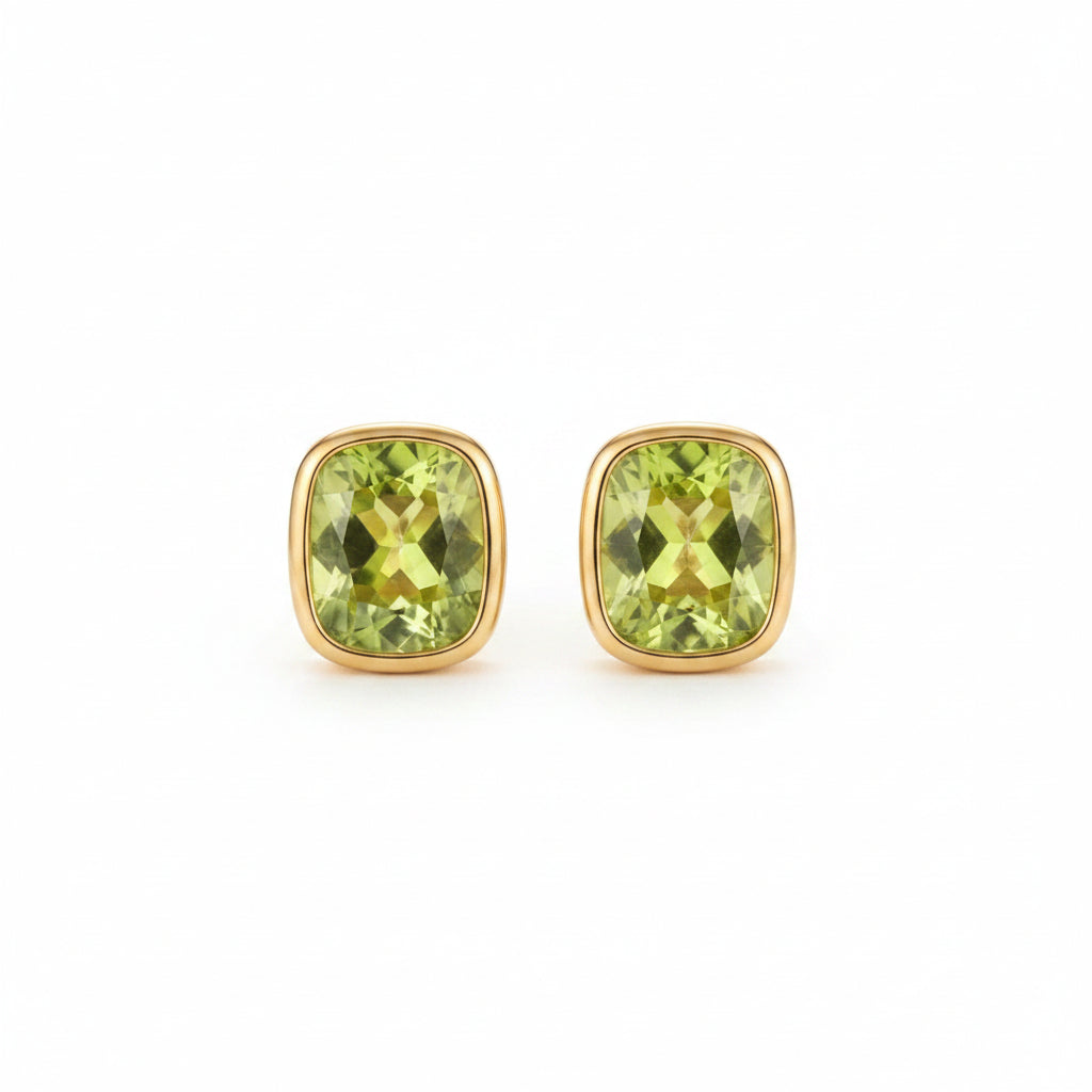 A pair of gold stud earrings with cushion-cut light green gemstones, resting on the pages of an open vintage book.