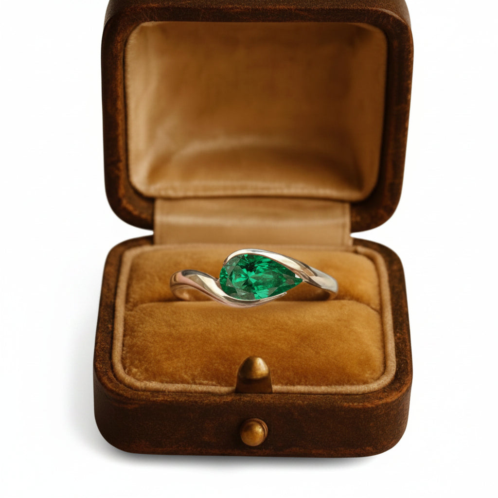 A silver ring with a pear-cut green emerald gemstone displayed in an open brown velvet jewelry box on a warm brown background.