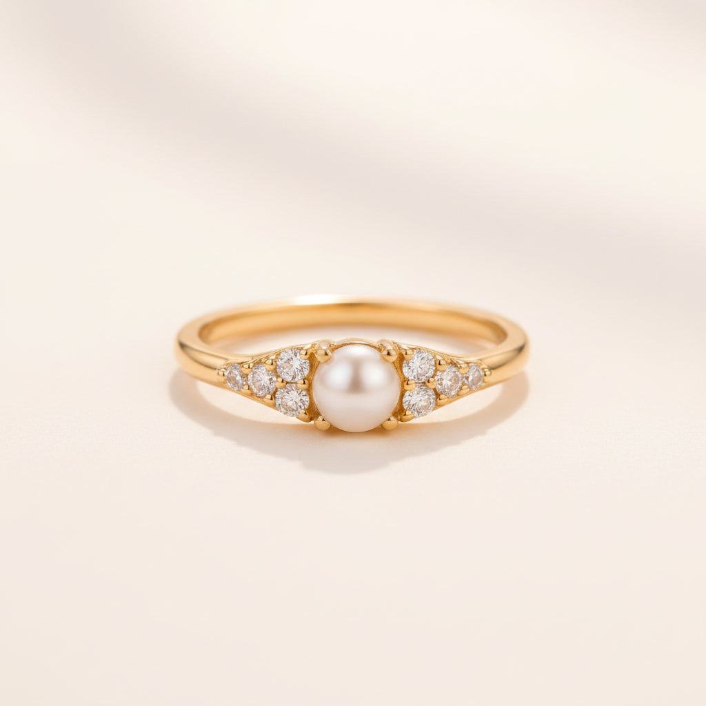 A gold ring with a central white pearl and small clear stones on either side, resting on a textured stone surface with a blurred green background.