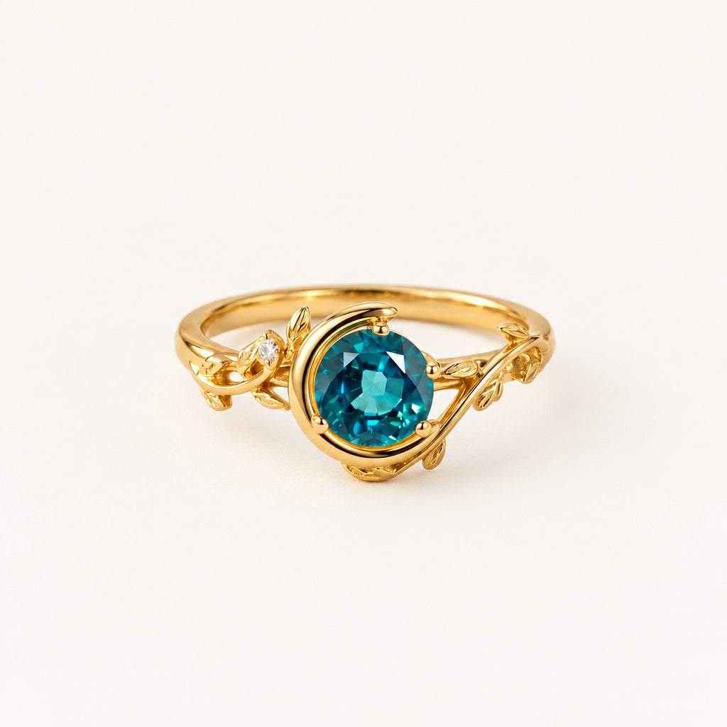 A gold ring with a round faceted teal blue gemstone, resting on a textured beige fabric with cream lace and dried floral elements in the background.