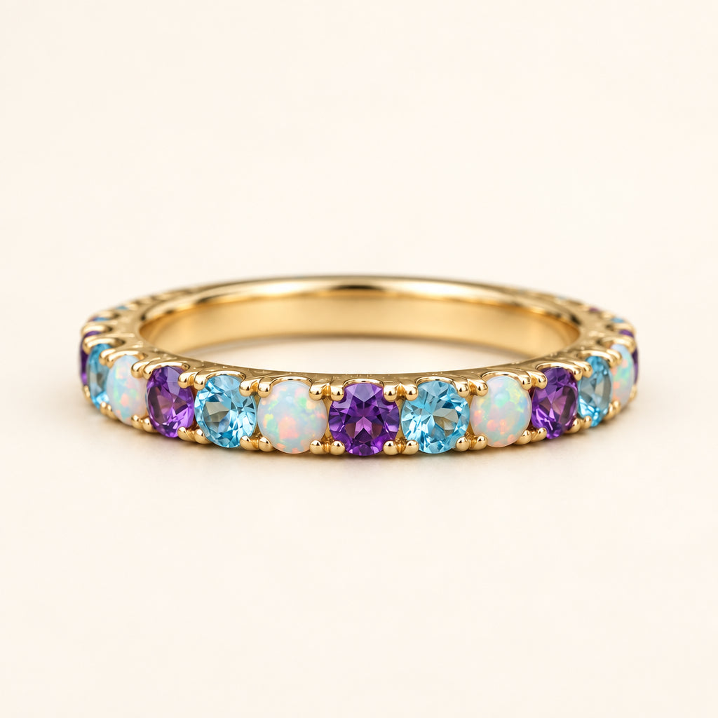 A gold eternity ring featuring a row of round-cut light blue topaz, purple amethyst, and white opal gemstones, resting on a textured stone surface with a blurred green background.