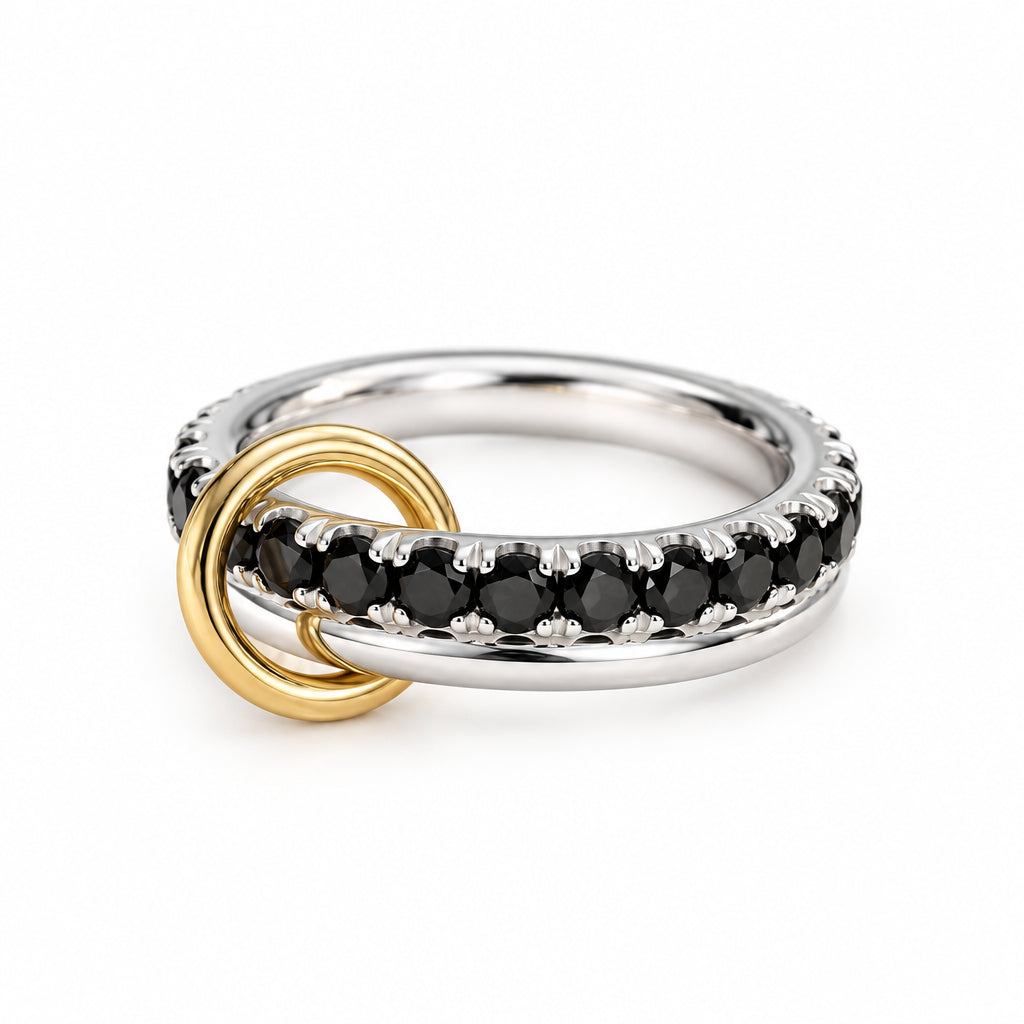 A close-up of three rings resting on textured cream fabric with soft green leaves blurred in the background. The rings include a polished yellow gold ring, a silver ring with small dark stones, and a silver ring with textured clear stones.