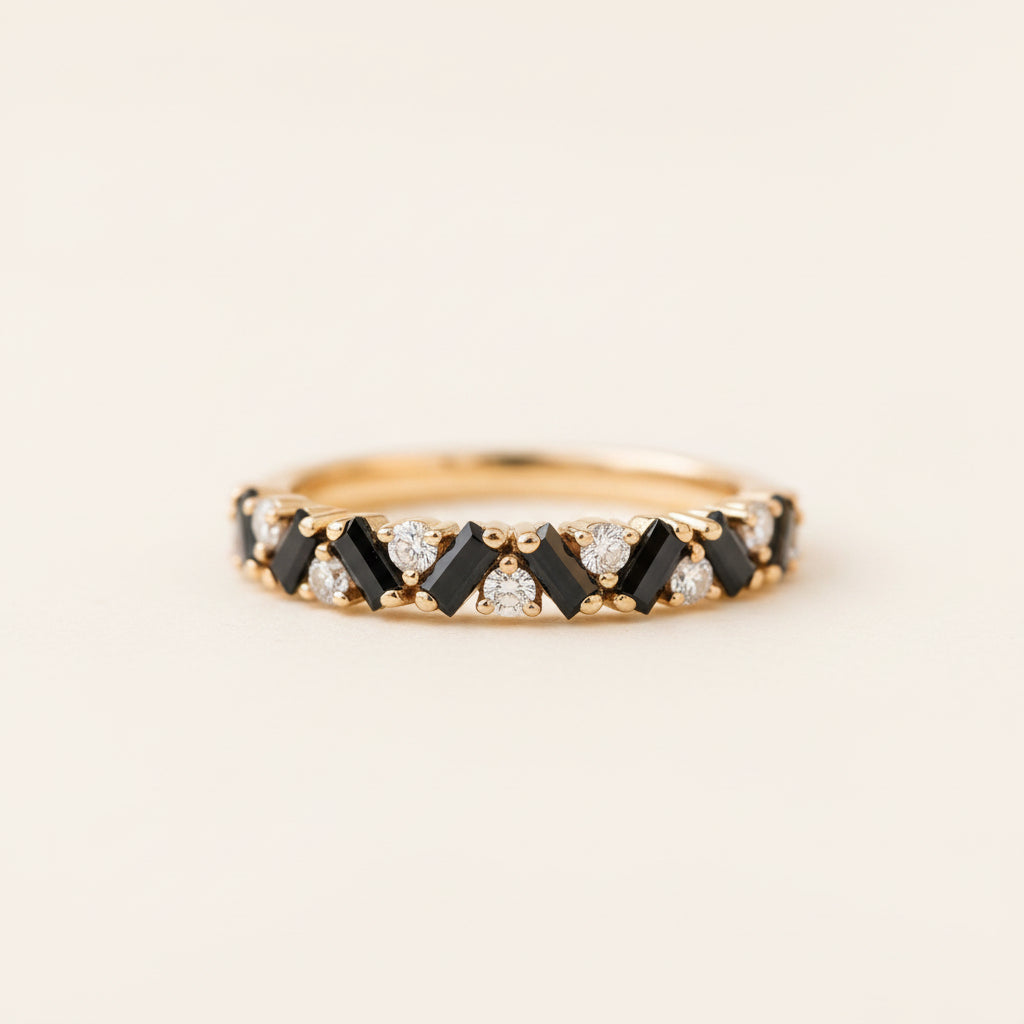A yellow gold eternity ring featuring alternating black baguette and round-cut clear gemstones, resting on a textured stone surface with a blurred green garden background.