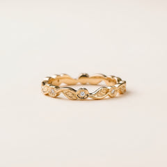 A delicate yellow gold eternity band ring with small round-cut diamonds set in a wavy, leaf-like pattern, resting on a textured stone surface with a blurred natural background.