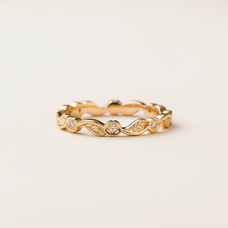 A delicate yellow gold eternity band ring with small round-cut diamonds set in a wavy, leaf-like pattern, resting on a textured stone surface with a blurred natural background.