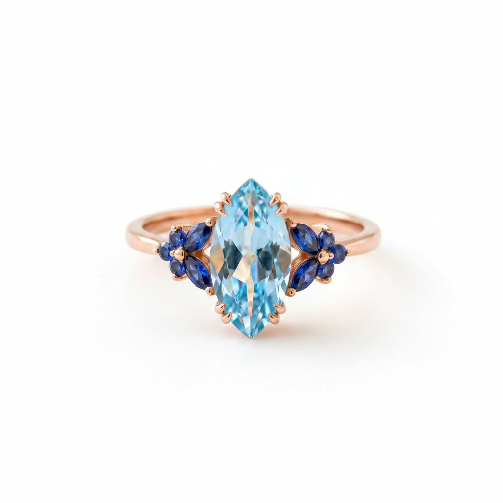 Gold ring with blue gemstone and floral accents on a textured surface with flowers.