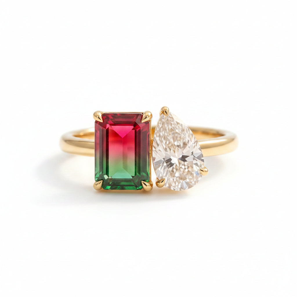 Gold ring with a red and green gemstone and a pear-shaped diamond on a textured surface with leaves in the background.