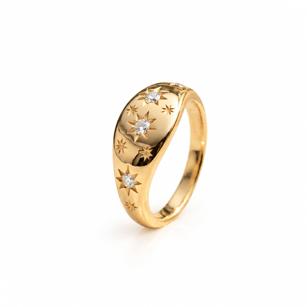 A gold ring with engraved star patterns and small clear gemstones, resting on a textured cream lace surface with soft-focus green leaves in the background.