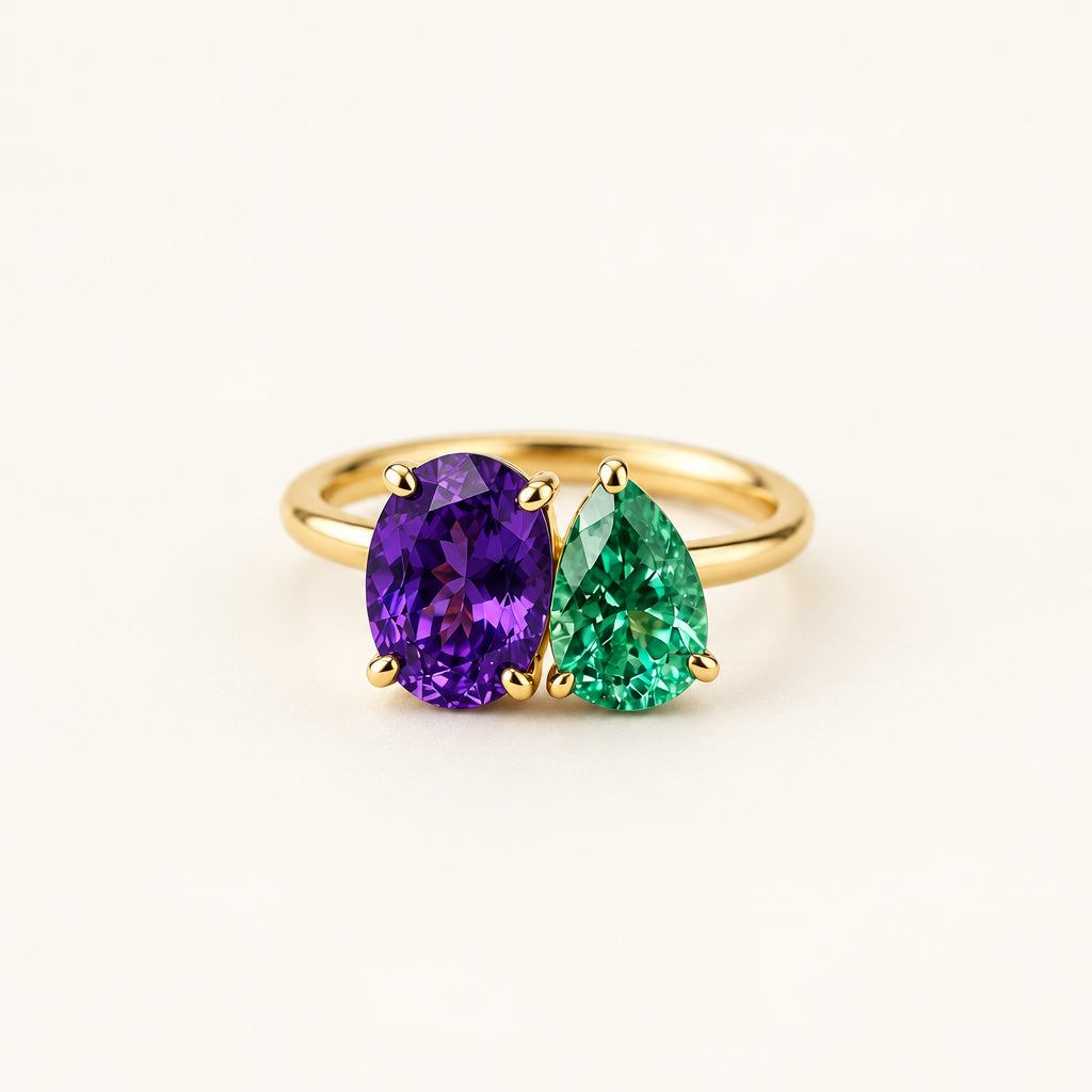 A gold ring featuring two marquise-cut gemstones, one deep purple and one emerald green, displayed on a textured beige fabric with dried flowers and lace.