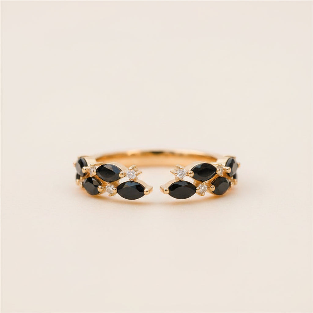 A gold ring with marquise-cut black gemstones and small clear accent stones, photographed resting on a textured stone surface with a softly blurred natural background.