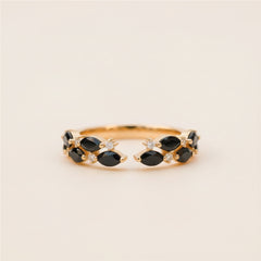 A gold ring with marquise-cut black gemstones and small clear accent stones, photographed resting on a textured stone surface with a softly blurred natural background.