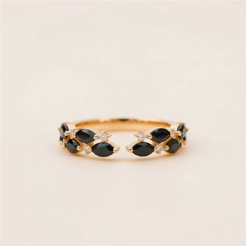A gold ring with marquise-cut black gemstones and small clear accent stones, photographed resting on a textured stone surface with a softly blurred natural background.