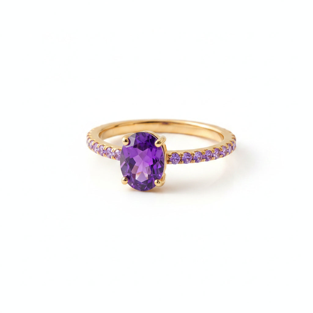A gold ring with a large oval purple gemstone and small purple accent stones along the band, displayed on a textured beige fabric with dried flowers.