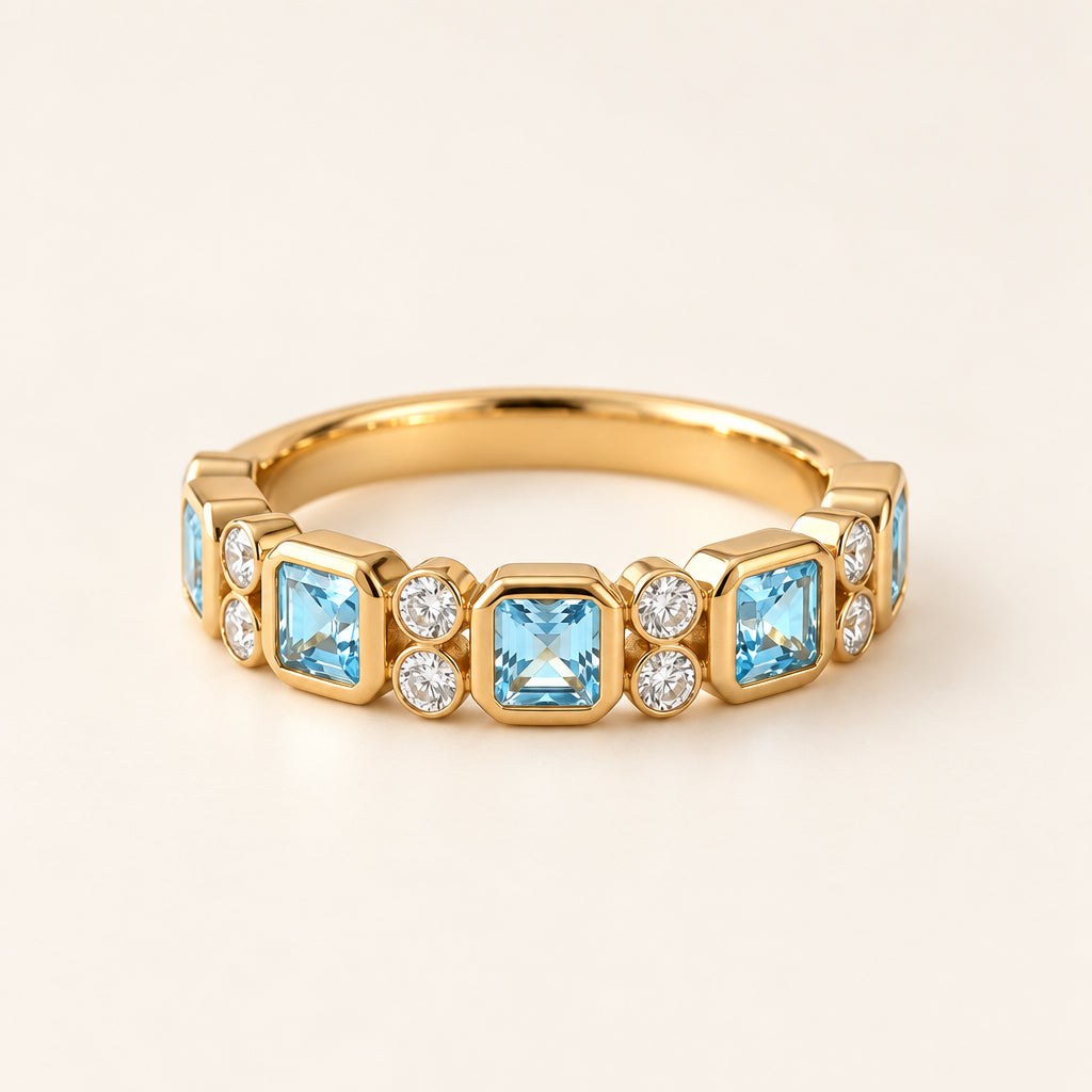 A gold band ring featuring five square-cut light blue gemstones and small clear round stones, styled on a beige fabric surface with lace and dried flowers.