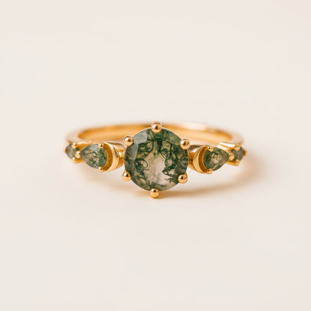 A gold ring with a central round green marbled gemstone and two smaller marquise-shaped green gemstones on either side, resting on a textured stone surface with a blurred natural background.