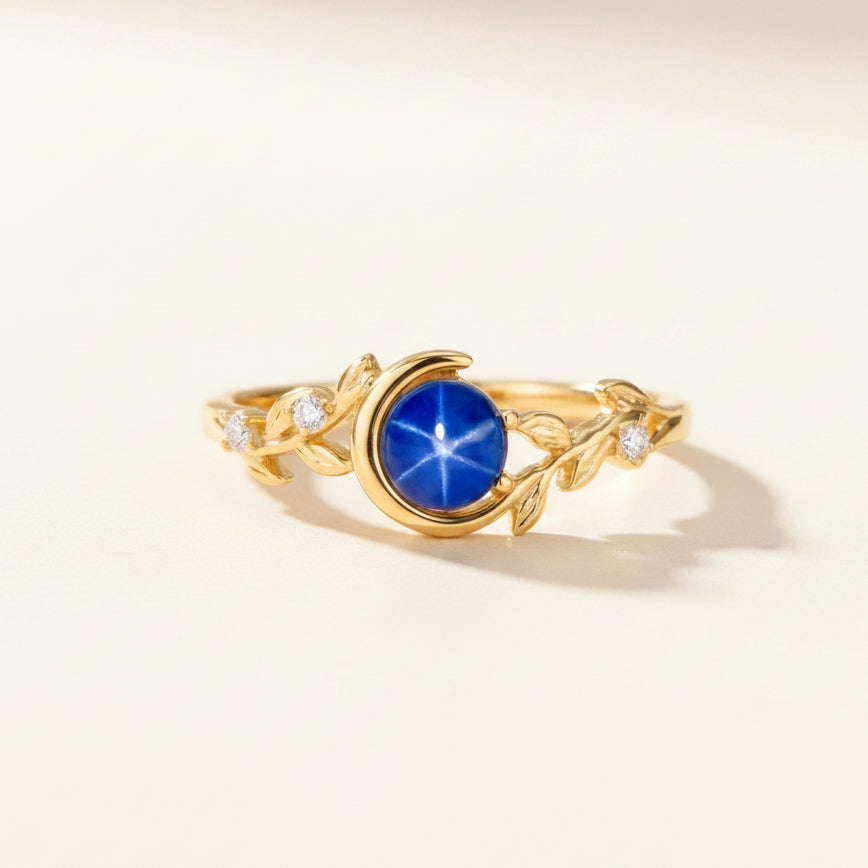 A gold ring featuring a central oval-cut deep blue star sapphire and delicate leaf-shaped side details, accented with small clear stones, displayed on a round gray stone pedestal.