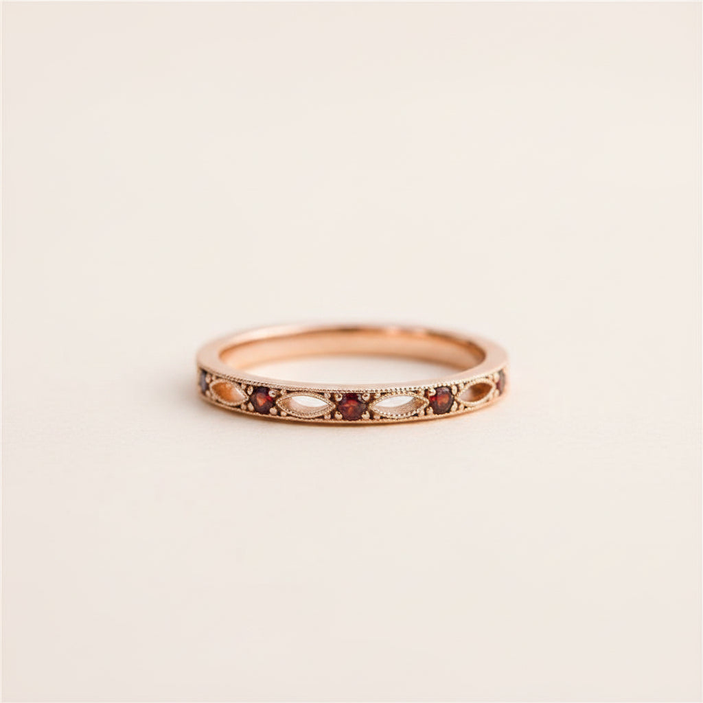 A rose gold eternity ring with small red garnet and white pearl accents, resting on a textured stone surface with a blurred green garden background.