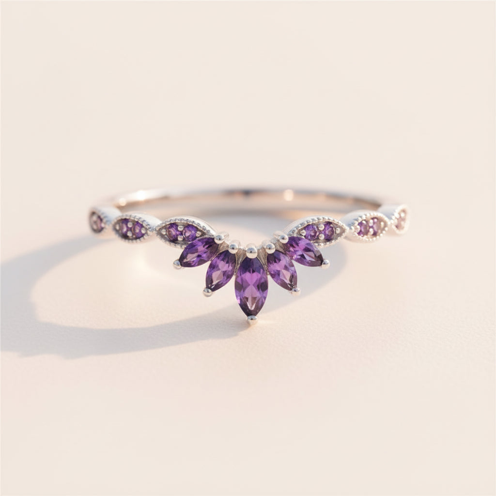 A silver-toned ring featuring a floral cluster of purple marquise-cut gemstones and small round purple stones, resting on a textured stone surface with a blurred natural background.