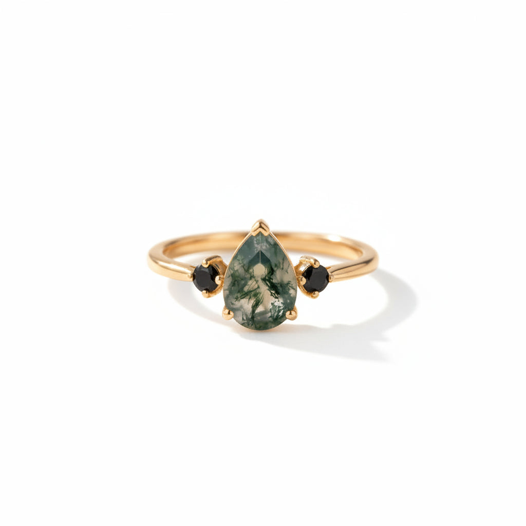 A gold ring with a teardrop-shaped green gemstone featuring white marbling, flanked by two small black round stones, resting on an old brown leather-bound book with lace in the background.