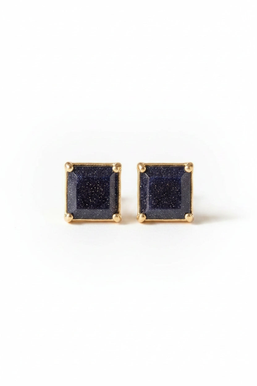 A pair of square stud earrings with dark blue glittery stones in gold settings, displayed on an antique brown book with lace in the foreground.