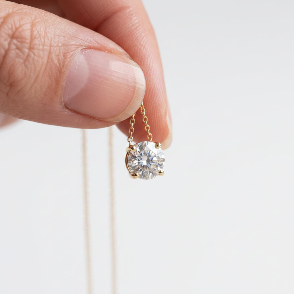 A close-up of a hand holding a delicate gold necklace with a round-cut clear gemstone pendant against a soft beige fabric background.