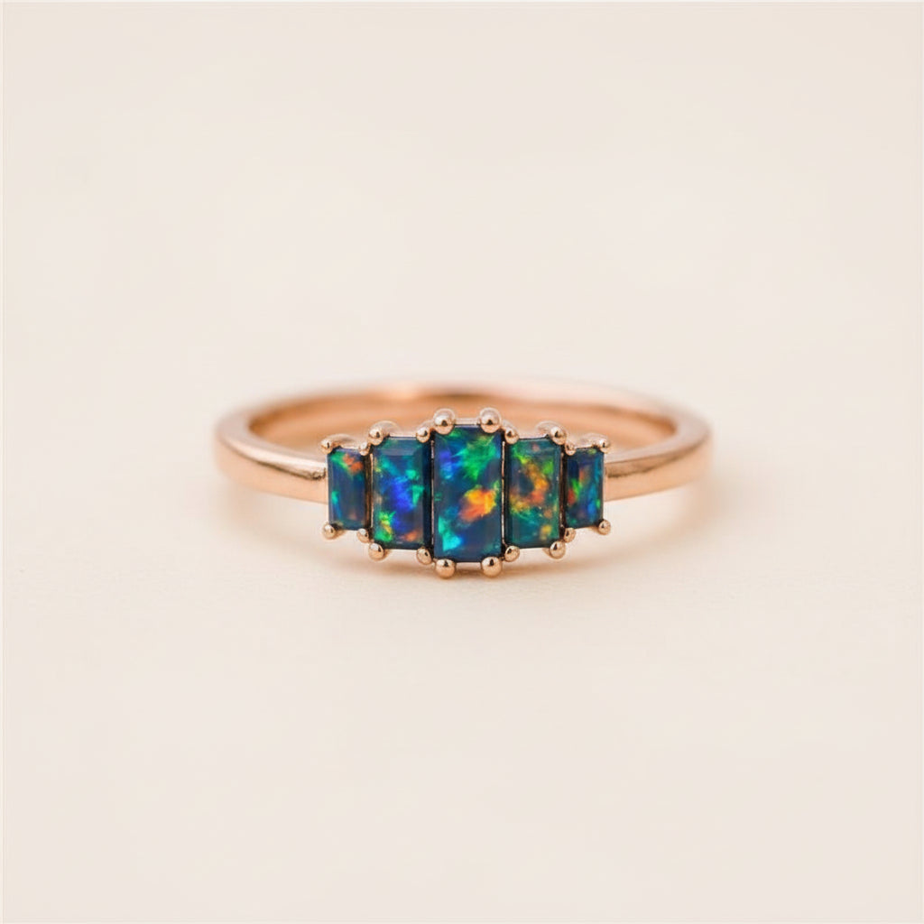 A five-stone ring featuring vibrant blue, green, and orange iridescent opal gemstones in a rose gold setting, resting on a textured stone surface.