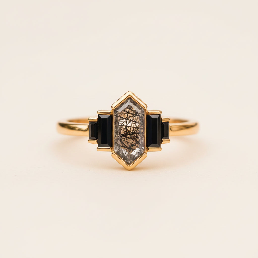A gold ring with a central clear crystal containing dark hair-like inclusions, flanked by two dark rectangular stones, resting on a textured stone surface with a blurred background.