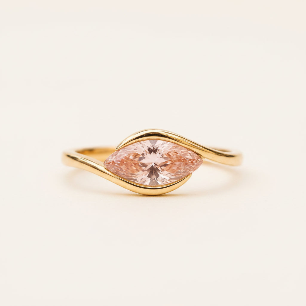 A gold ring with a marquise-cut peach-pink gemstone, resting on a textured stone surface with a blurred natural background.