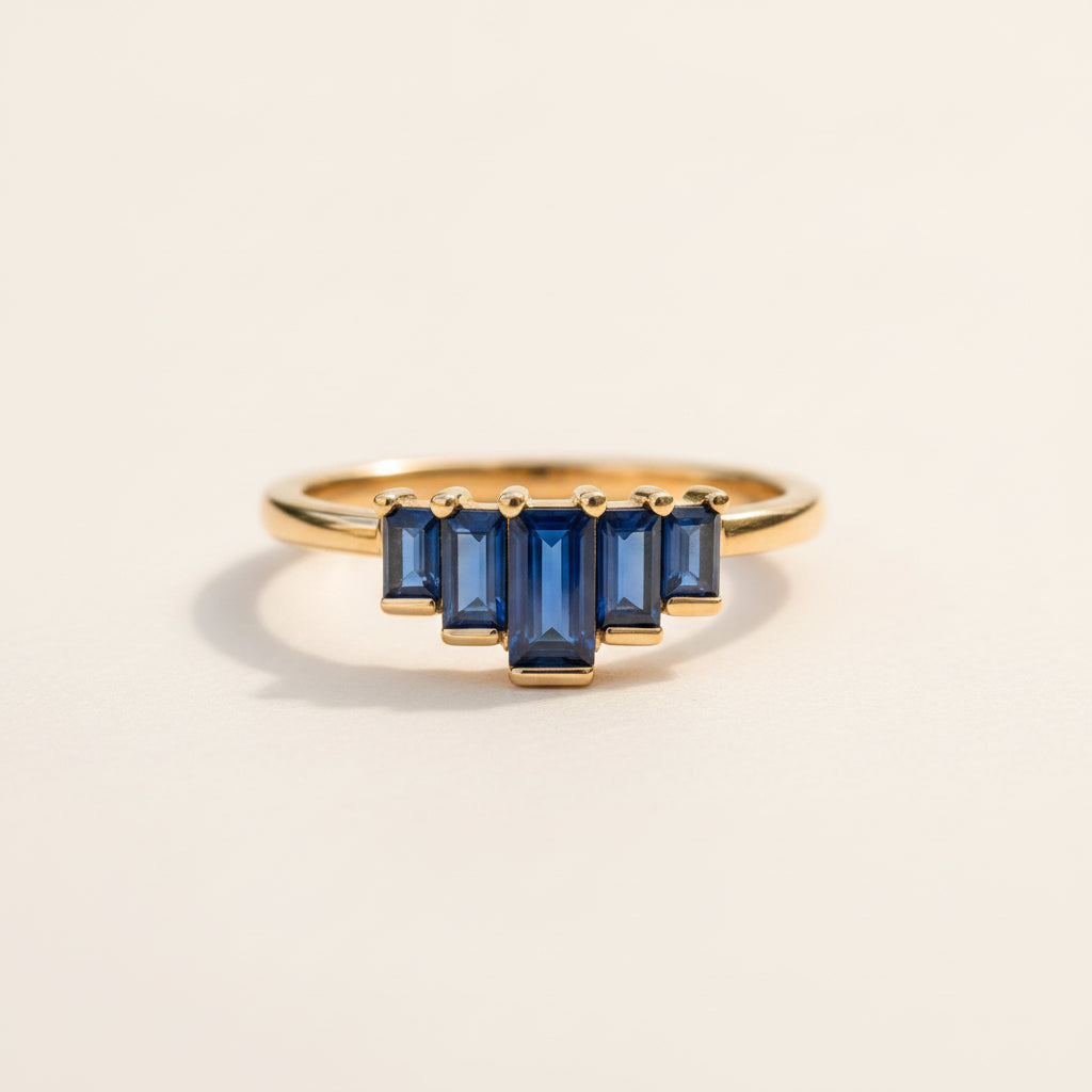 A gold ring featuring a row of five baguette-cut deep blue sapphires, displayed on a textured stone with a soft-focus green background.