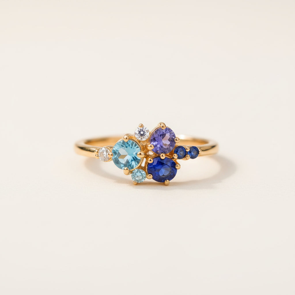 A gold ring with a floral cluster of blue and light blue gemstones, resting on a textured wooden surface with a blurred green background.