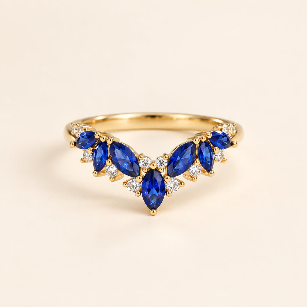 Gold V-shaped ring with marquise-cut blue sapphires and small clear accent stones, displayed on textured cream fabric with soft green leaves blurred in the background.
