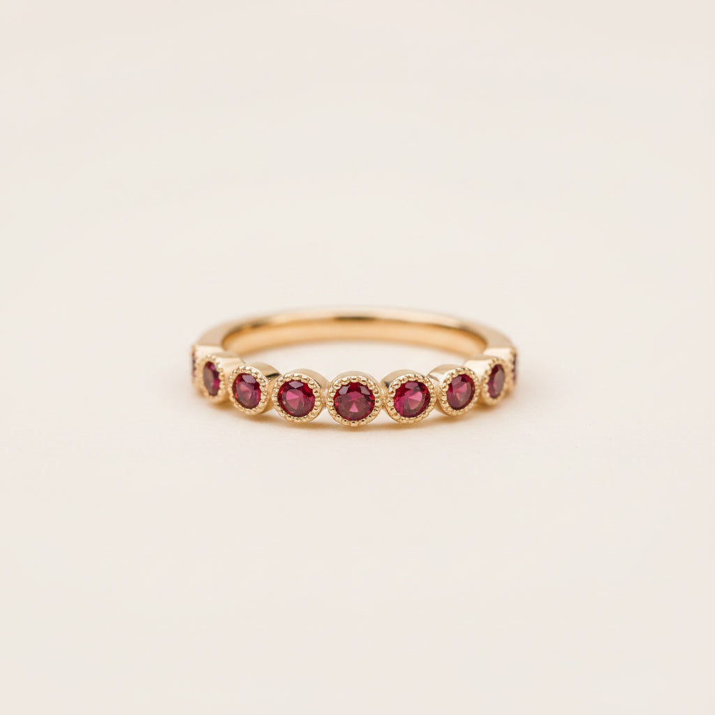 A gold eternity ring featuring round red gemstones, photographed resting on a textured stone surface with a soft, blurred natural background.
