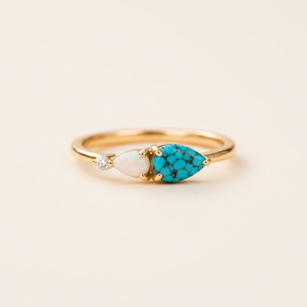 A gold ring featuring a marquise-cut turquoise stone, a small oval white opal, and a tiny clear gemstone, resting on a textured stone surface with a blurred natural background.