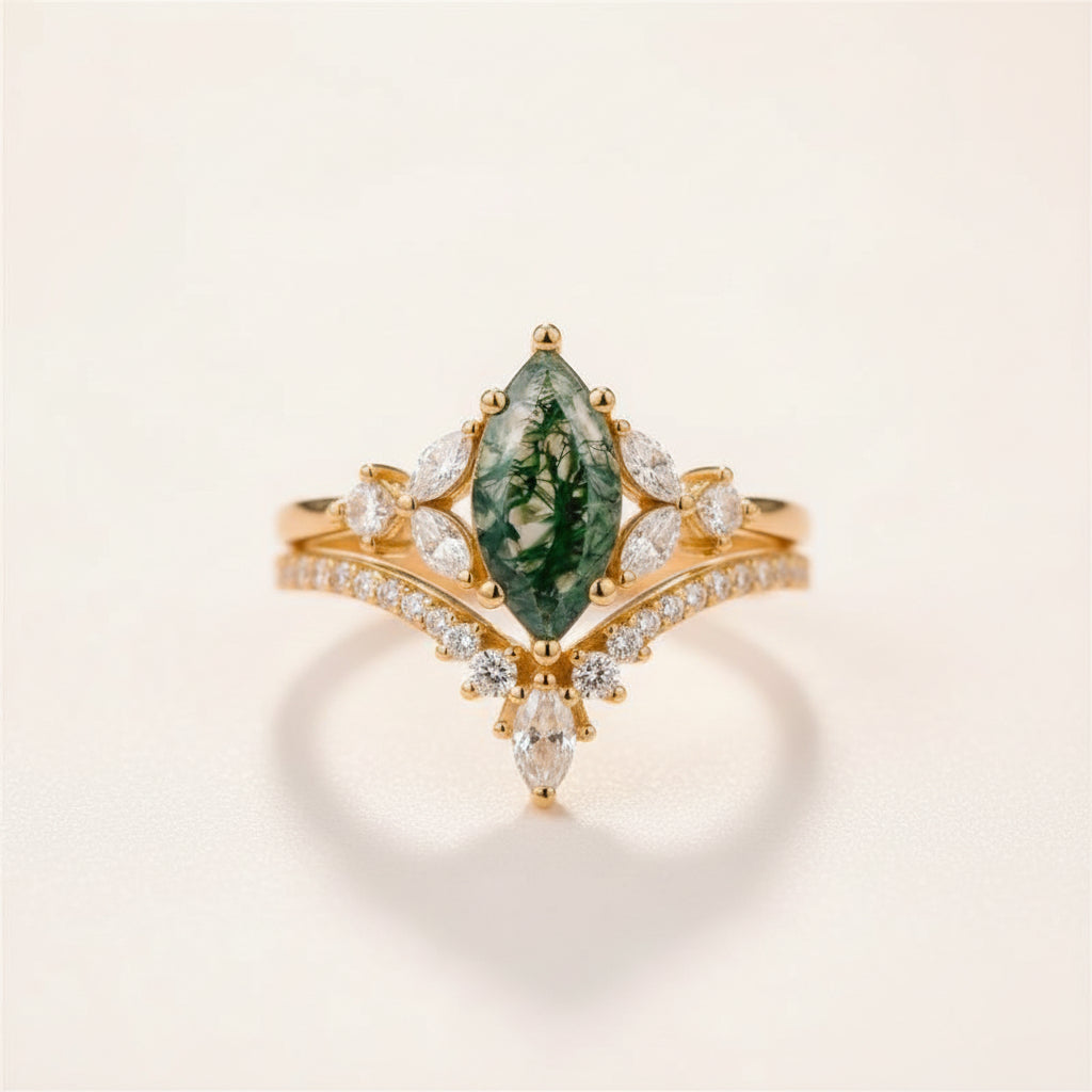 A gold ring with a marquise-cut green forest gemstone and small clear stones, resting on a textured stone surface with a blurred natural background.