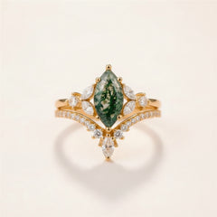 A gold ring with a marquise-cut green forest gemstone and small clear stones, resting on a textured stone surface with a blurred natural background.