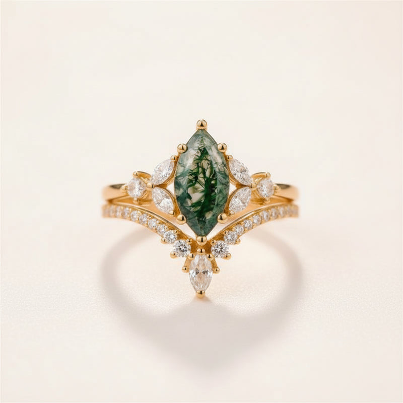 A gold ring with a marquise-cut green forest gemstone and small clear stones, resting on a textured stone surface with a blurred natural background.