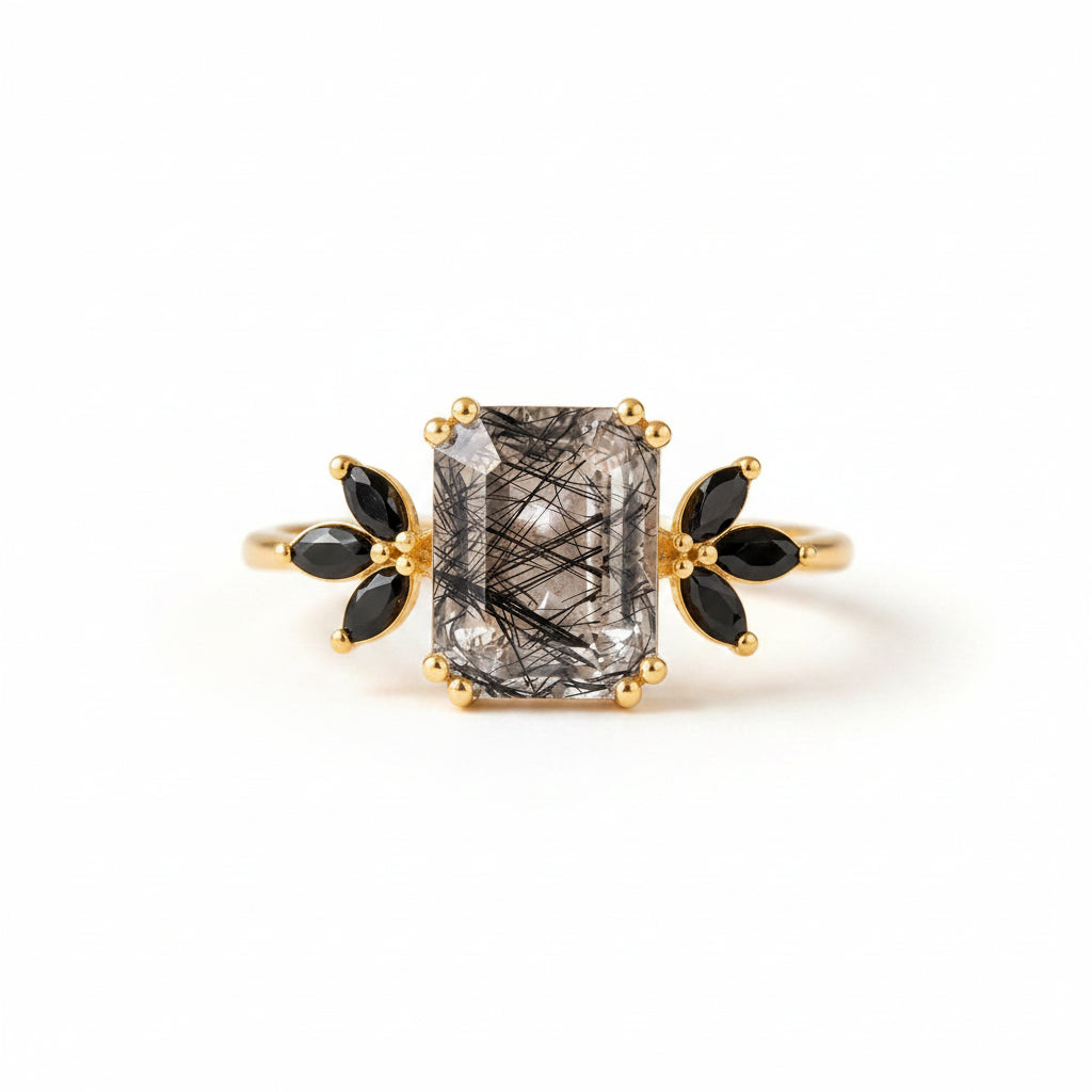 A gold ring featuring a rectangular clear gemstone with black needle-like inclusions, flanked by black marquise-shaped stones, displayed on an antique book with lace.