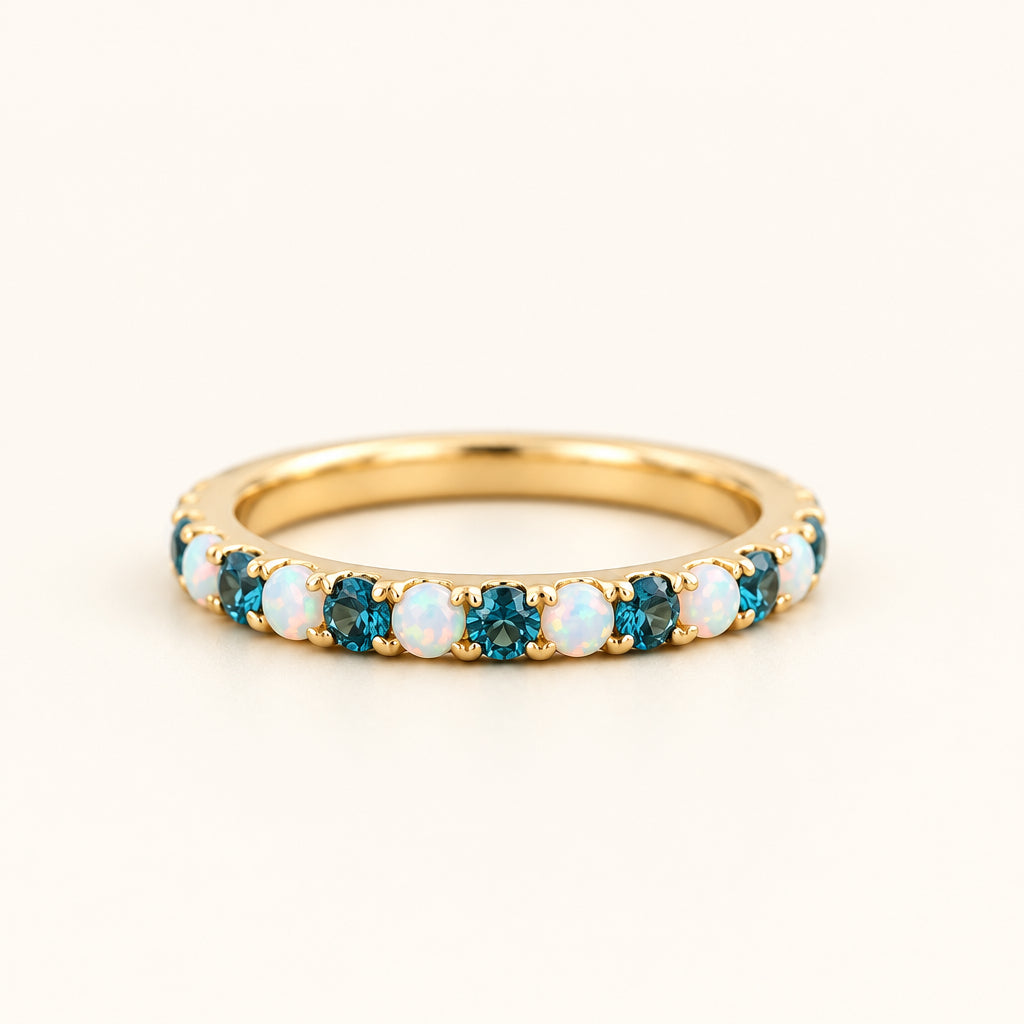 A gold eternity ring featuring alternating round-cut white opal and deep blue topaz gemstones, resting on a textured stone surface with a soft-focus natural background.
