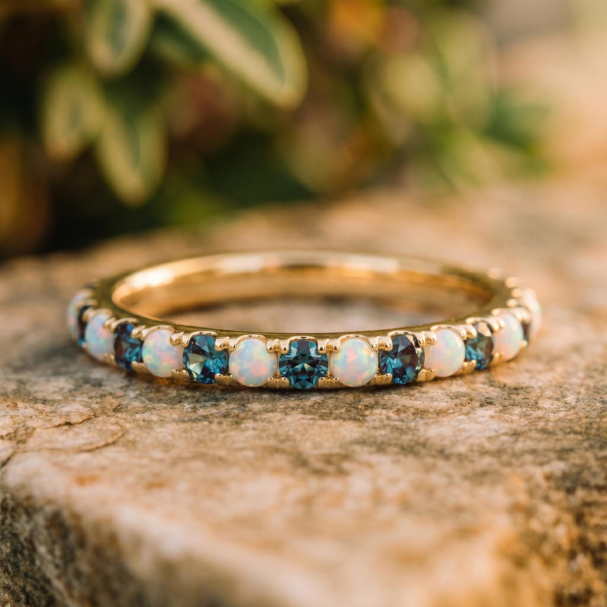 A gold eternity ring featuring alternating round-cut white opal and deep blue topaz gemstones, resting on a textured stone surface with a soft-focus natural background.