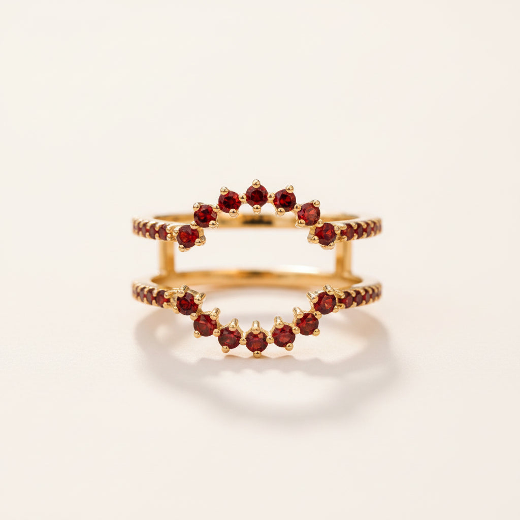 A pair of gold rings featuring a wavy band set with small round red gemstones, resting on a textured stone surface with a blurred natural background.
