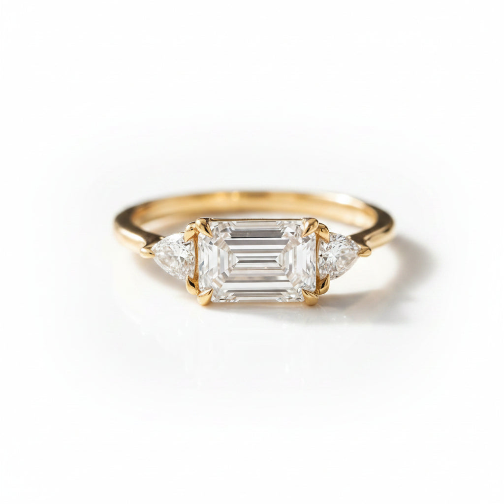 A yellow gold three-stone ring featuring a central emerald-cut diamond flanked by two smaller round diamonds, resting on a dark wooden surface.