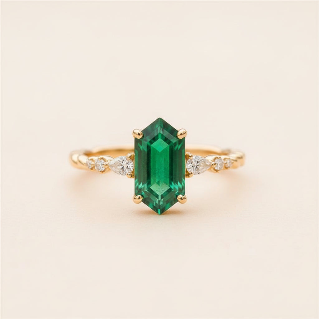 A gold ring featuring a large emerald-cut green gemstone center with small clear accent stones on the band, resting on a textured stone surface with a blurred natural background.