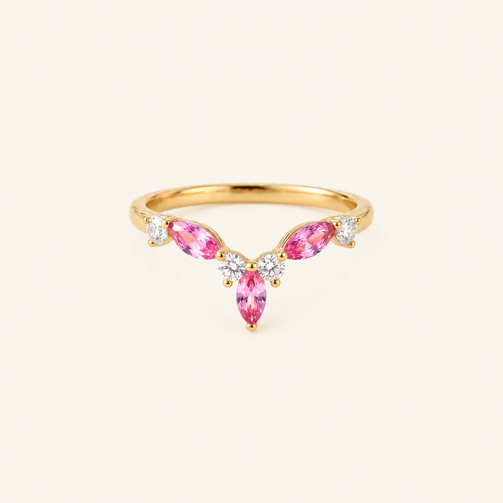A gold ring featuring three marquise-cut pink gemstones in a V-shape setting, accented with small clear round stones, displayed on a textured beige fabric with lace and dried flowers.
